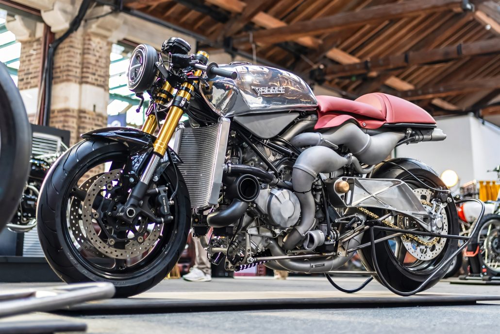 Veloce Aperion 2026 – Linke Frontansicht auf Motorradmesse mit poliertem Aluminiumtank, rotem Ledersitz, goldenen Öhlins-Gabeln und freiliegendem X8-Zweitaktmotor mit Resonanzauspuffkrümmern
