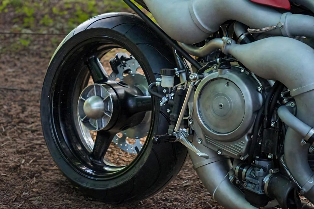 Veloce Aperion 2026 – Hinterrad- und Motordetail mit Pullrod-Hinterradfederung, Getriebedeckel, Expansionskammern und Bremsscheibe auf Waldboden