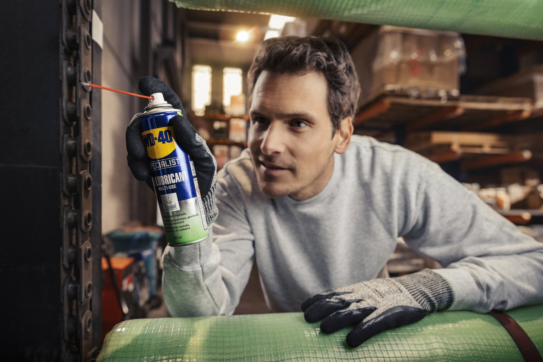Mann mit Arbeitshandschuhen sprüht WD-40 Specialist Bio-Schmierspray Multifunktional über das Sprühröhrchen auf ein Metallteil in einer Lagerwerkstatt