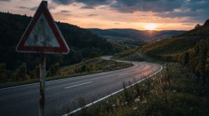 Kurvige Landstrasse durch herbstliche Weinberge bei Sonnenuntergang, im unscharfen Vordergrund ein rot-weisses Gefahrenwarnschild ohne lesbare Beschriftung, leere Fahrbahn, Symbolbild zur Gefahr auf beliebten Motorradstrecken