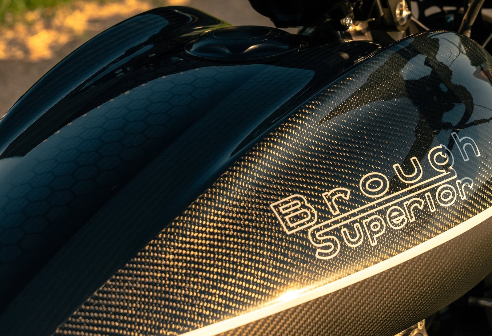 Brough Superior Dagger S: Was die neue Sportvariante wirklich ändert 3 Brough Superior Dagger S 2026 – Tank-Detailansicht bei natürlichem Licht im Freien, Carbon-Oberfläche mit goldenem Schimmer und Waben-Struktur, Brough Superior Schriftzug in Gold, Tankdeckel