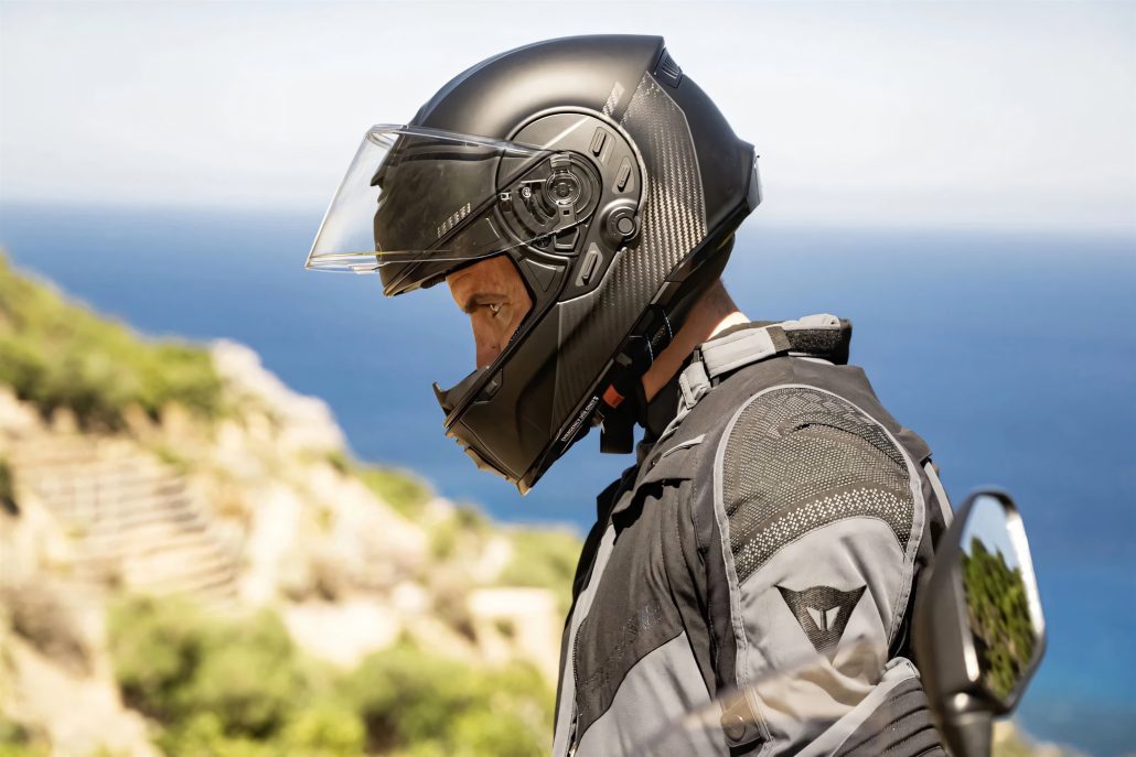 Cardo Beyond GTS gewinnt Red Dot Award vor dem Marktstart 5 Motorradfahrer mit geöffnetem Visier trägt den schwarzen Cardo Beyond GTS Helm mit integrierter Kommunikationseinheit vor Küstenkulisse mit Meer und Felsen