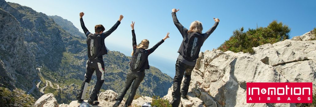 In&motion Emergency Call: Airbag ruft jetzt auch in Deutschland Hilfe 5 Drei Motorradfahrer in schwarzer Schutzkleidung mit sichtbaren In&motion-Airbag-Westen stehen auf einem Felsen mit Blick auf Berge und Meer und strecken jubelnd die Arme in die Höhe, unten rechts das In&motion-Airbags-Logo