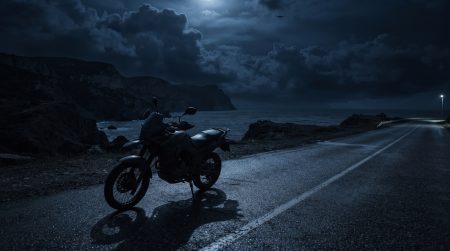 Nachtaufnahme eines geparkten Reise-Motorrads an einer einsamen Kuestenstrasse auf der Krim, im Hintergrund das Schwarze Meer und Steilkuesten unter dramatischem Wolkenhimmel, am Himmel die Silhouette einer kleinen Drohne, KI-generiertes Symbolbild zum naechtlichen Motorrad-Fahrverbot.