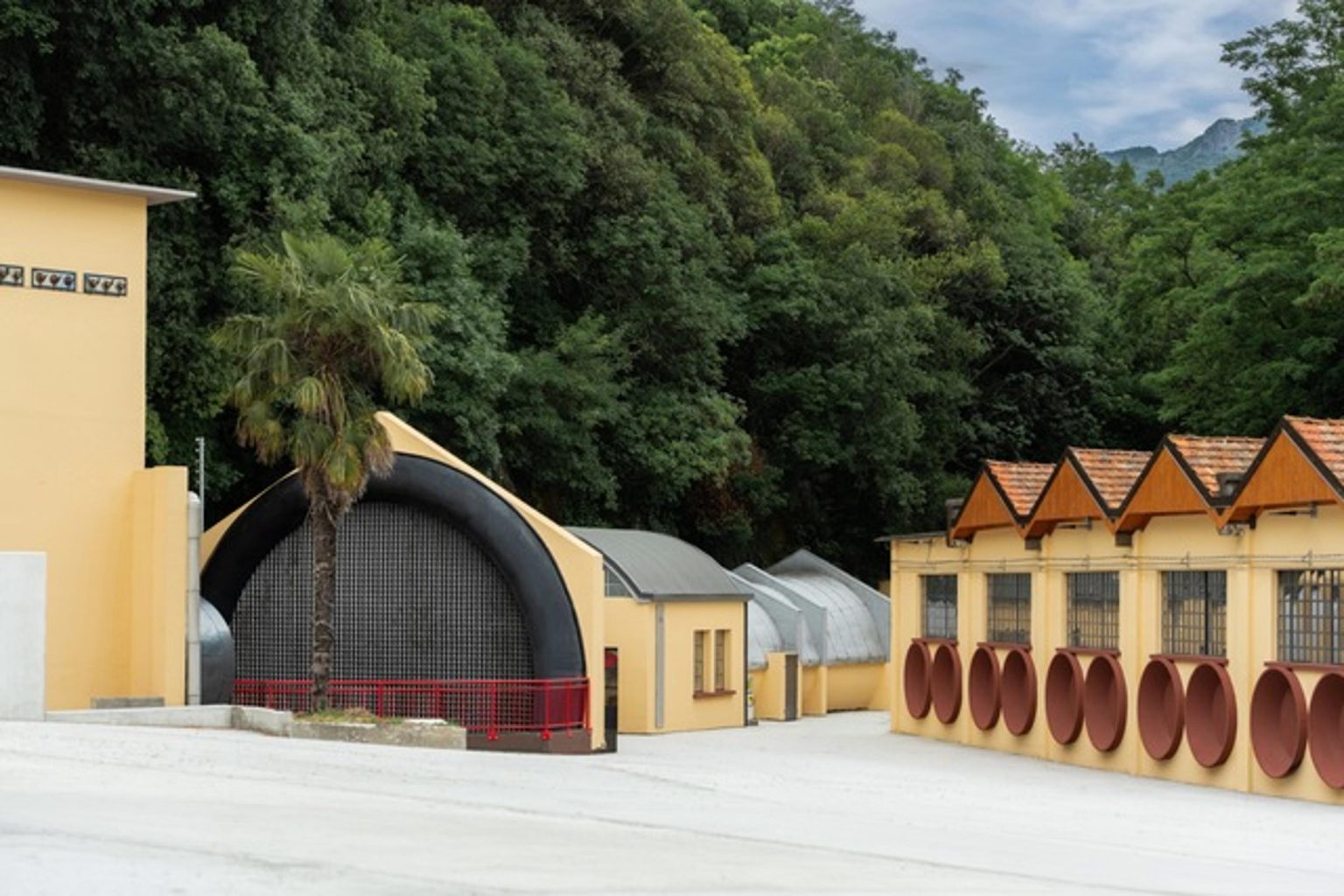 Moto Guzzi World Days 2026 – Gesamtansicht des neuen Werksgeländes in Mandello del Lario mit Palme, rundem Hallenbau und Bergpanorama am Comer See