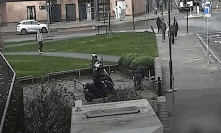 Körnige Überwachungskamera-Aufnahme von oben: ein abgestellter Motorroller auf einem Grünstreifen an einer Straßenkreuzung in Bristol, im Hintergrund Passanten und parkende Autos