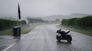 KI-generiertes Symbolbild: einsames Rennmotorrad auf leerer, regennasser Straßenrennstrecke im Nebel, schwarze Flagge am Streckenrand, leere Tribüne im Hintergrund – stiller, pietätvoller Moment ohne Personen
