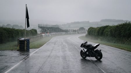 KI-generiertes Symbolbild: einsames Rennmotorrad auf leerer, regennasser Straßenrennstrecke im Nebel, schwarze Flagge am Streckenrand, leere Tribüne im Hintergrund – stiller, pietätvoller Moment ohne Personen