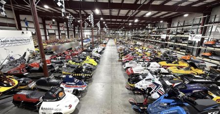 Dutzende bunt lackierte Renn-Schneemobile mit Startnummern stehen in langen Reihen in einer Lagerhalle mit Stahldach, im Hintergrund ein Schild der Apple Auctioneering Co. und karierte Zielflaggen unter der Decke, links ein einzelnes Custom-Motorrad.