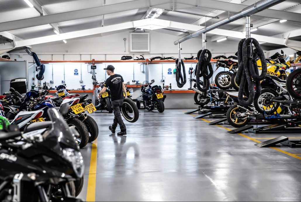 SuperBike Factory ist insolvent: Das bedeutet es für Motorradfahrer 6 Werkstattbereich von SuperBike Factory mit Mitarbeiter, der einen Reifen trägt, umgeben von aufgebockten und geparkten Gebrauchtmotorrädern verschiedener Marken