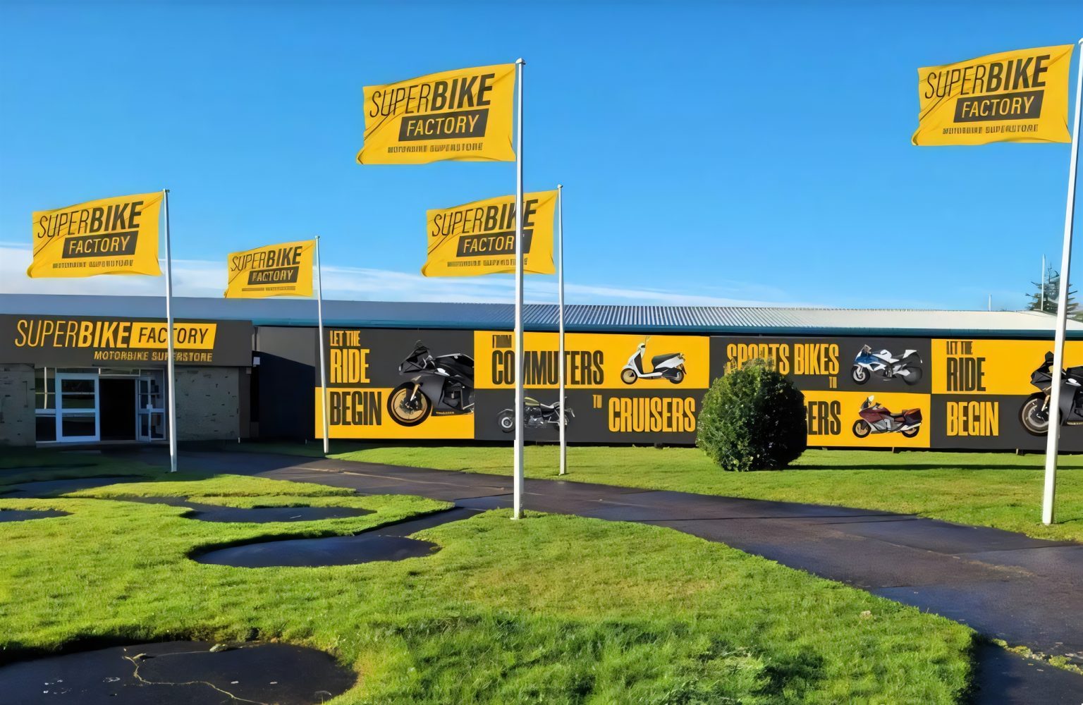 Außenansicht eines SuperBike-Factory-Gebäudes mit mehreren gelben Fahnen und großflächiger Wandwerbung 'Let The Ride Begin' vor blauem Himmel