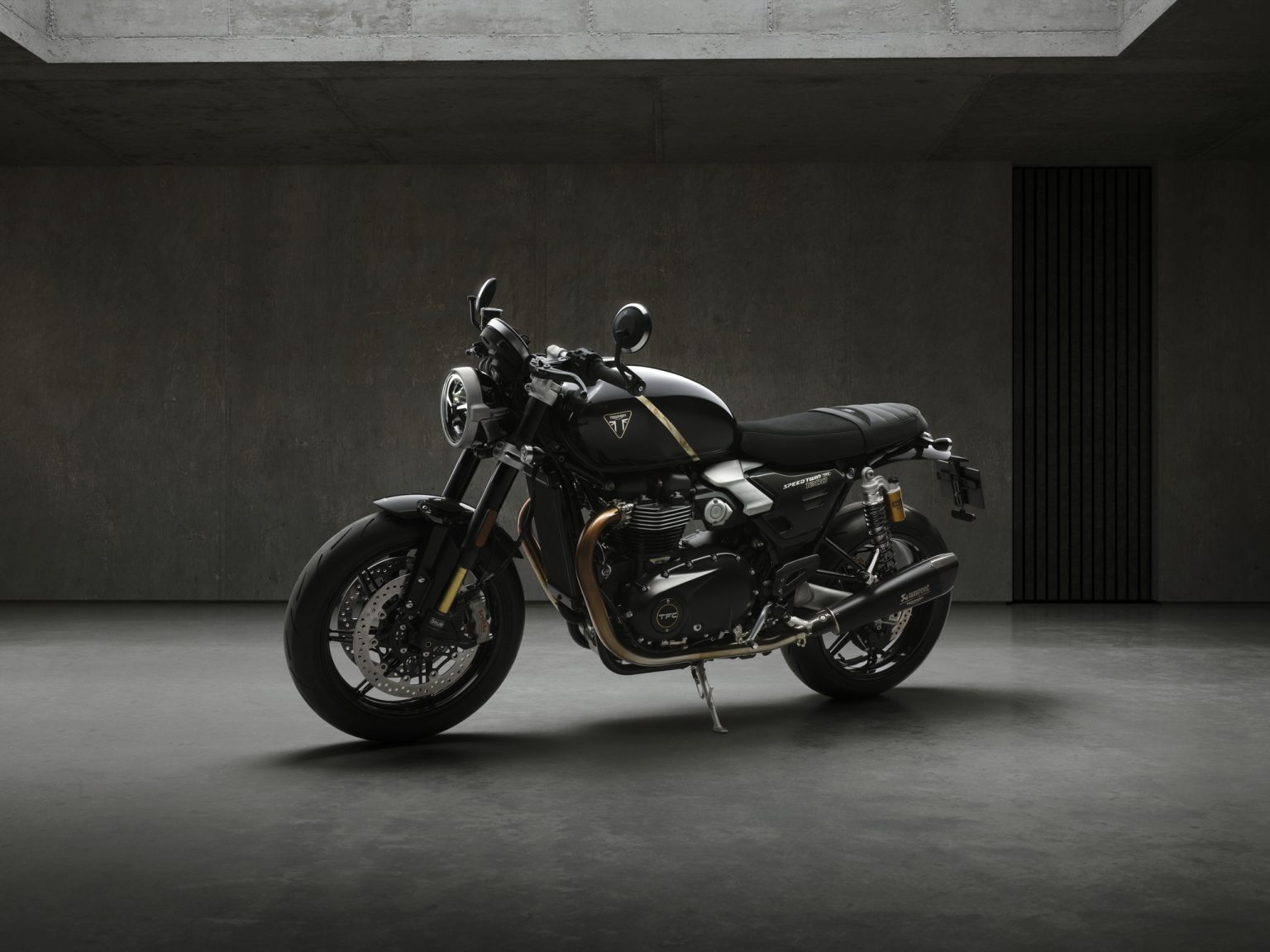 Triumph Speed Twin 1200 TFC: 750er-Sonderserie mit Öhlins und Akrapovič Triumph Speed Twin 1200 TFC 2027 – linke Frontperspektive im Betonstudio, Gesamtansicht in Obsidian Gold mit Öhlins-Gabel und Akrapovič-Schalldämpfer