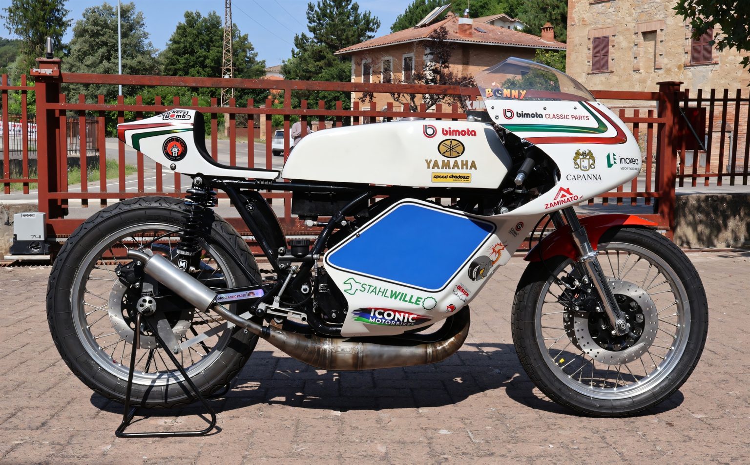 Yamaha/Bimota TZ 350 GP 2026 – Seitenansicht von links in Weiß mit Italien-Streifen, rotem Kotflügel und blauem Startnummernfeld, auf Pflaster vor rotem Zaun in der Toskana