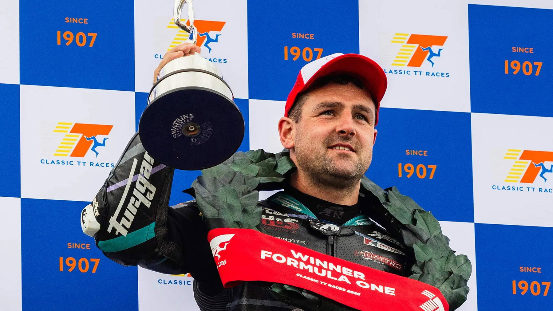 Classic TT: Dunlop Breaks the Anstey Record and Wins Twice in a Day 4 Michael Dunlop reckt mit roter Siegermütze, Lorbeerkranz und Schärpe Winner Formula One den Siegerpokal vor der Classic-TT-Sponsorenwand in die Höhe – Isle of Man 2026