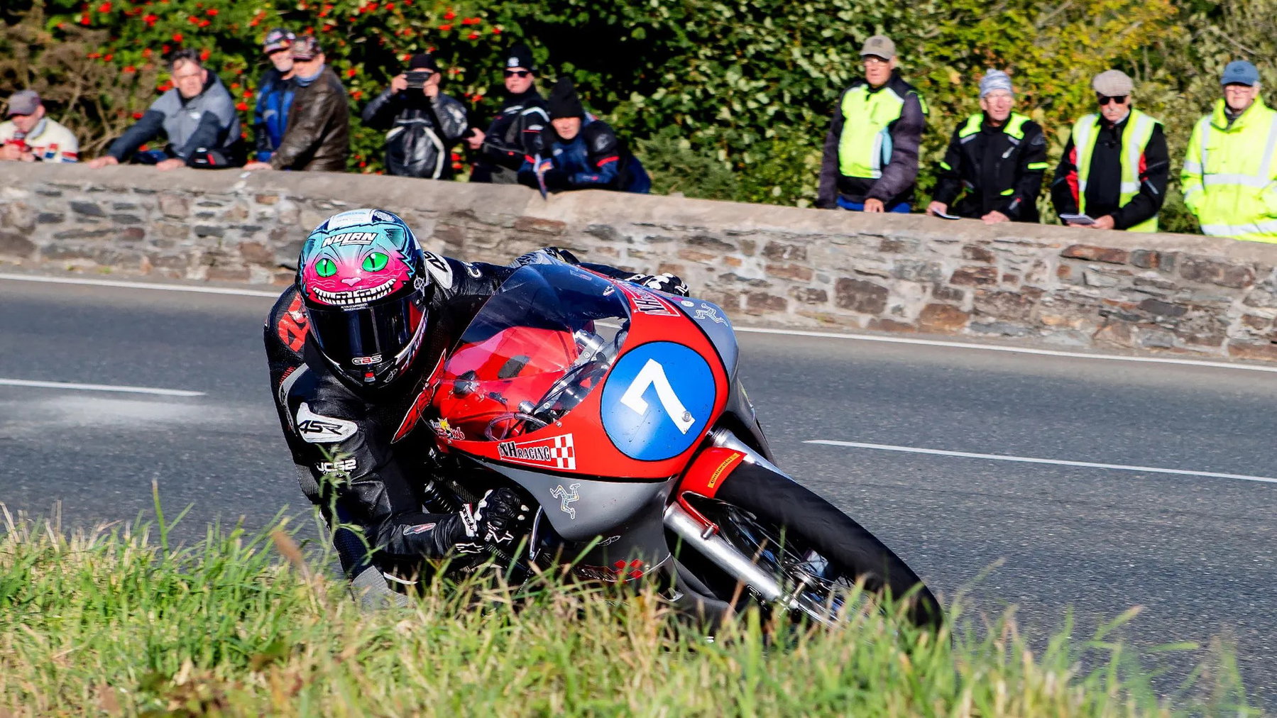 Dominic Herbertson in tiefer Schräglage auf der rot-silbernen Nummer 7 von Nigel Haith Racing, dahinter Zuschauer an der Steinmauer – Historic Junior Classic TT, Isle of Man 2026