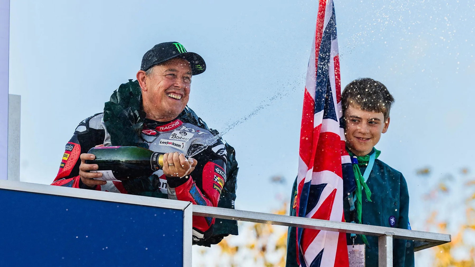 John McGuinness versprüht mit Lorbeerkranz und schwarzer Kappe Champagner auf dem Podium, daneben ein junger Fahnenträger – Historic Junior Classic TT, Isle of Man 2026