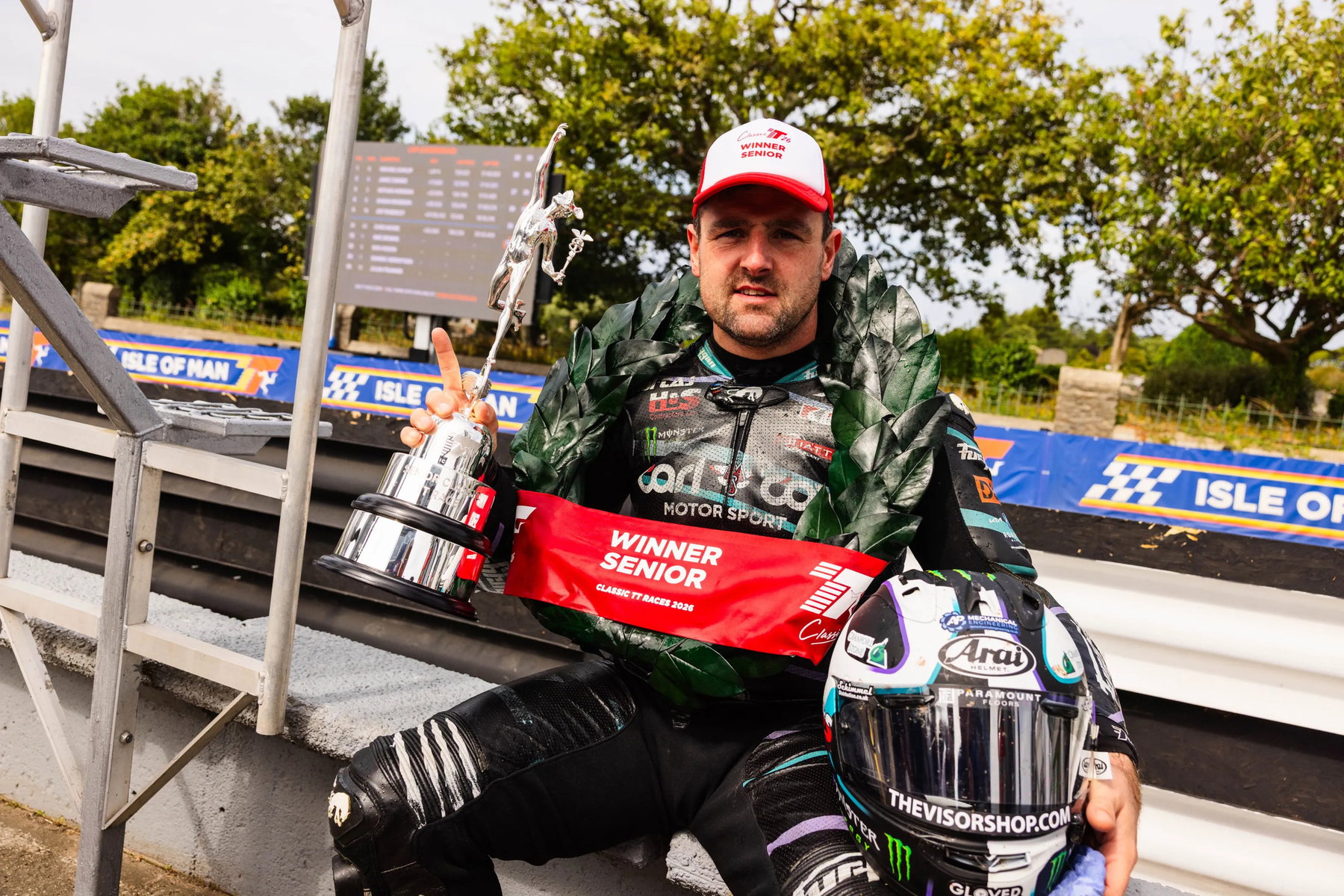 Michael Dunlop sitzt mit Siegerpokal, Lorbeerkranz und roter Winner-Senior-Schärpe an der Boxenmauer und hebt den Zeigefinger – Senior Classic TT, Isle of Man 2026