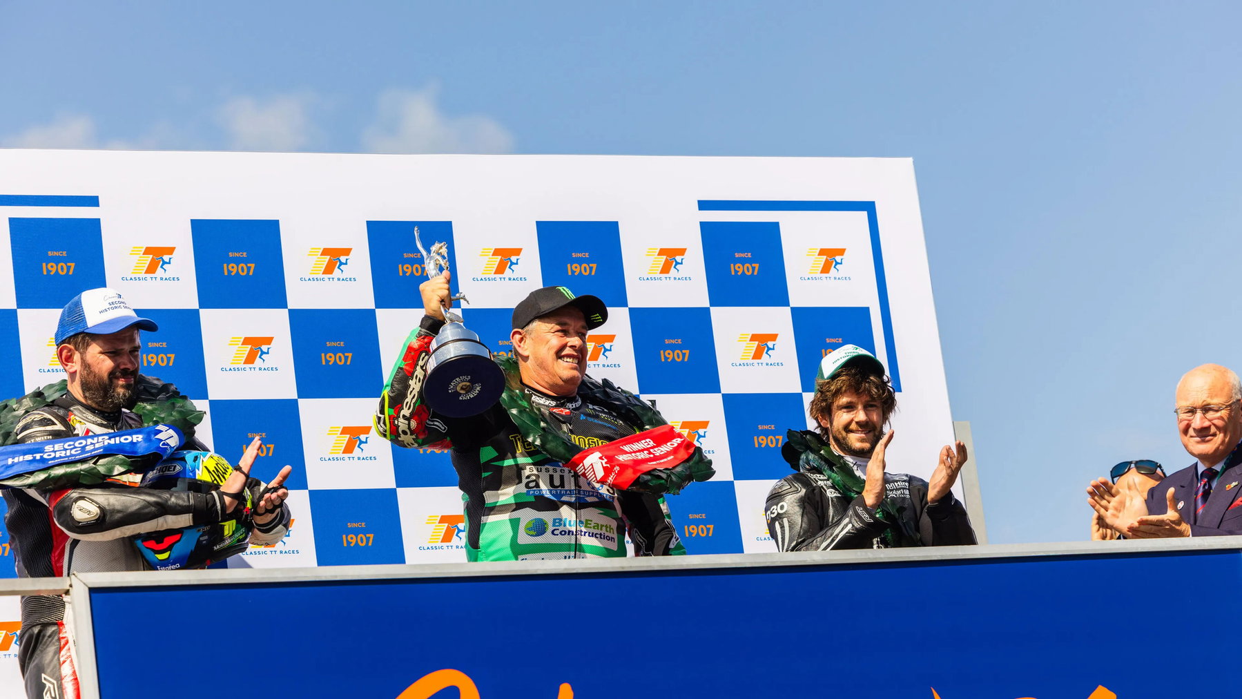 McGuinness gewinnt Historic Senior: erster Sieg seit sieben Jahren 6 John McGuinness reckt auf dem Podium den Pokal in die Höhe, links Shaun Anderson mit Zweiter-Platz-Schärpe, rechts Mike Browne – Historic Senior, Classic TT 2026, Isle of Man