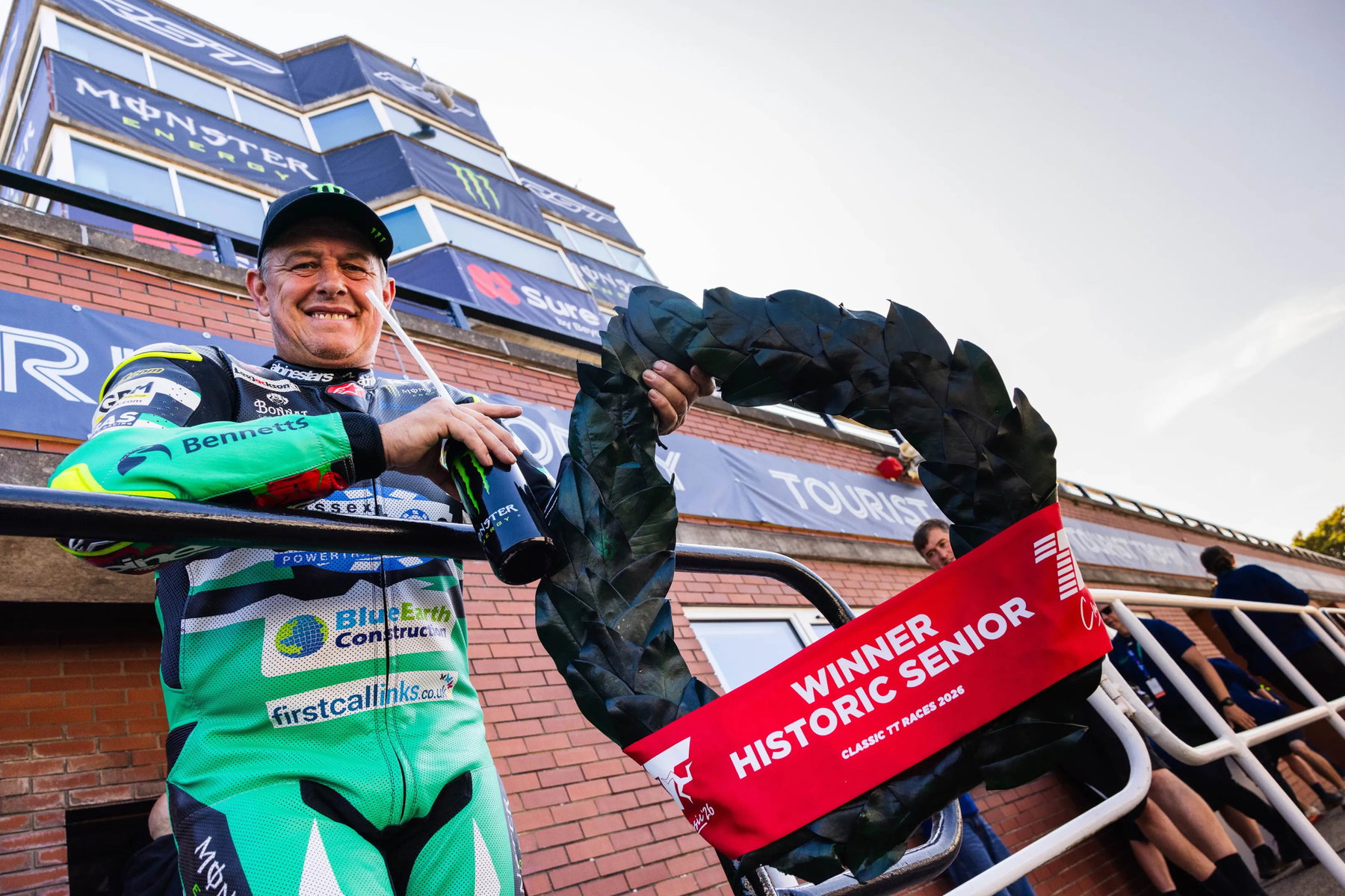 McGuinness gewinnt Historic Senior: erster Sieg seit sieben Jahren 3 John McGuinness mit Siegerkranz und der roten Schärpe Winner Historic Senior vor dem Grandstand-Gebäude – Classic TT 2026, Isle of Man