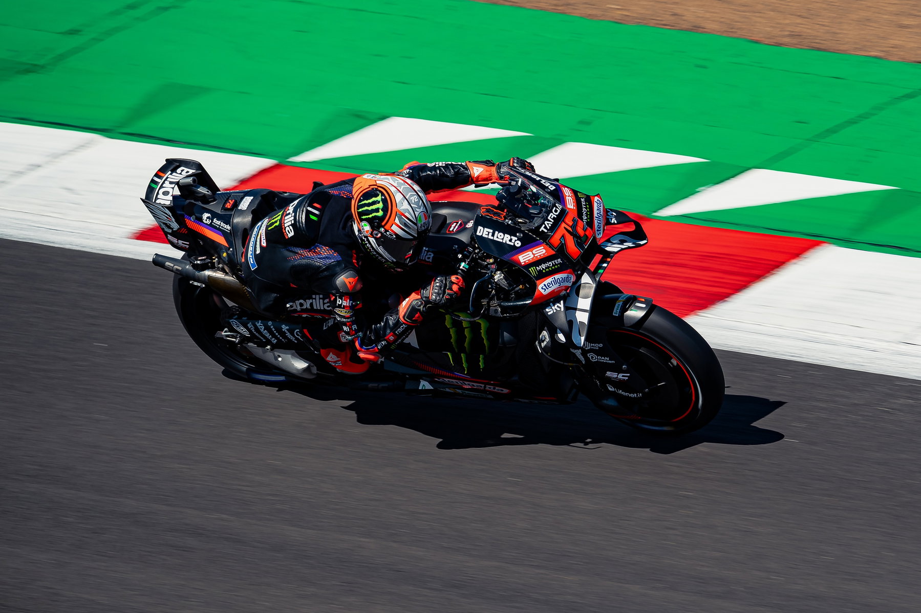 Aprilia holt in Silverstone alle drei Podestplätze: Marquez nur Neunter 5 Marco Bezzecchi aus der Vogelperspektive in Schräglage auf der schwarzen Aprilia RS-GP über dem grün-roten Randstein – MotoGP Großbritannien GP Silverstone 2026