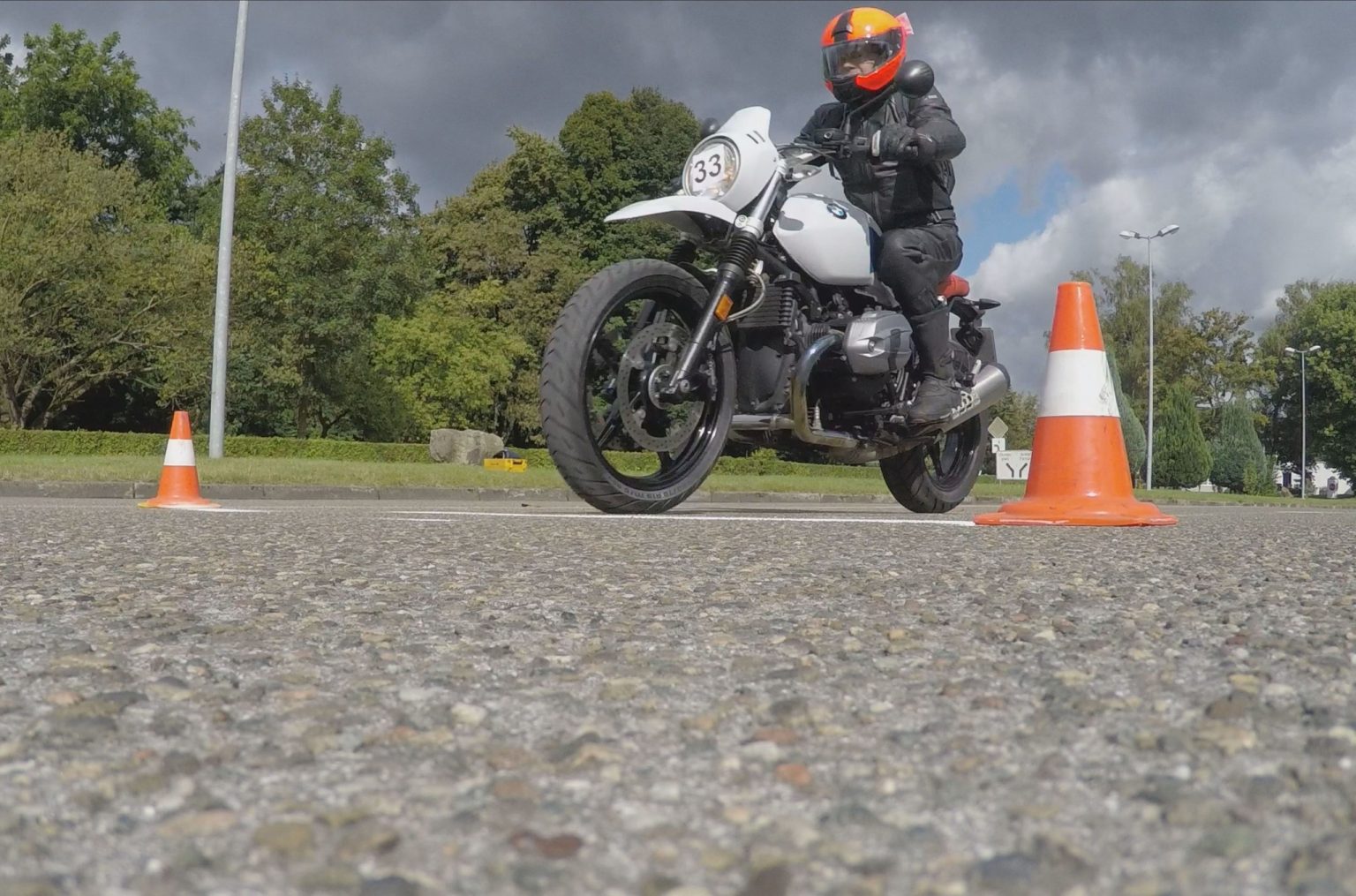 Motorradfahrer in schwarzer Lederkombi mit orangefarbenem Helm auf weißem Retro-Motorrad mit Startnummer 33 bremst bei einem Fahrversuch zwischen zwei orange-weißen Pylonen auf einem Übungsplatz, Froschperspektive, im Hintergrund Bäume und bewölkter Himmel