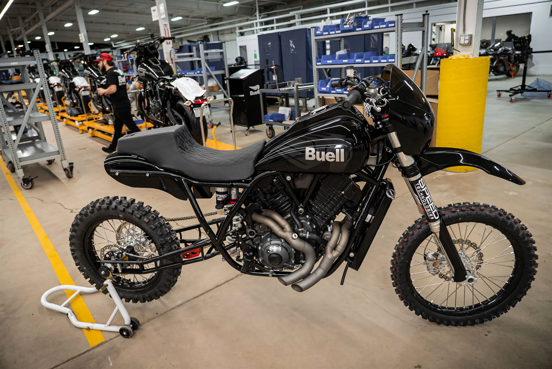 Buell The Goat Bergrenn-Konzept 2026 – Seitenansicht des kompletten Umbaus in der Fertigungshalle, im Hintergrund weitere Buell-Prototypen und ein Mitarbeiter, deutlich sichtbar der zweiflutige, seitlich austretende Auspuff.