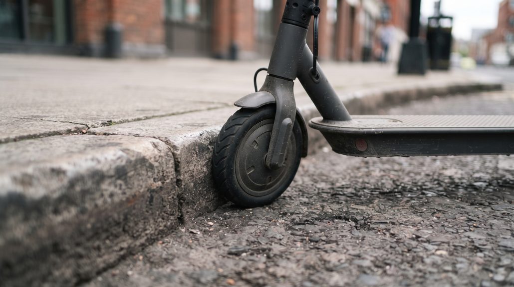 E-Scooter-Studie: Hirnverletzungen häufiger als bei Motorradfahrern 5 Nahaufnahme des kleinen, ungefederten Vorderrads eines E-Scooters direkt an einer Bordsteinkante als typische Sturzursache