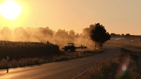 Grüner Traktor mit Anbaugerät am Feldrand direkt neben einer Landstraße, dichte Staubwolke im Gegenlicht der untergehenden Sonne über der Fahrbahn