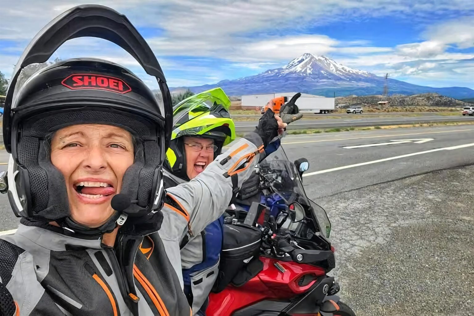 Zwei Motorradfahrerinnen in Reisemontur machen ein Selfie auf einer Landstraße, im Hintergrund ein schneebedeckter Vulkan