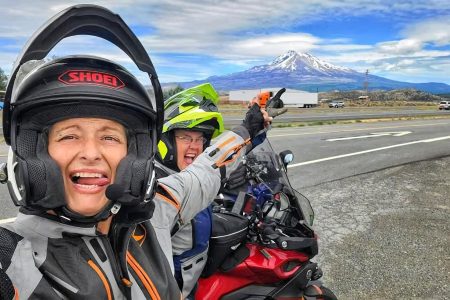 Zwei Motorradfahrerinnen in Reisemontur machen ein Selfie auf einer Landstraße, im Hintergrund ein schneebedeckter Vulkan