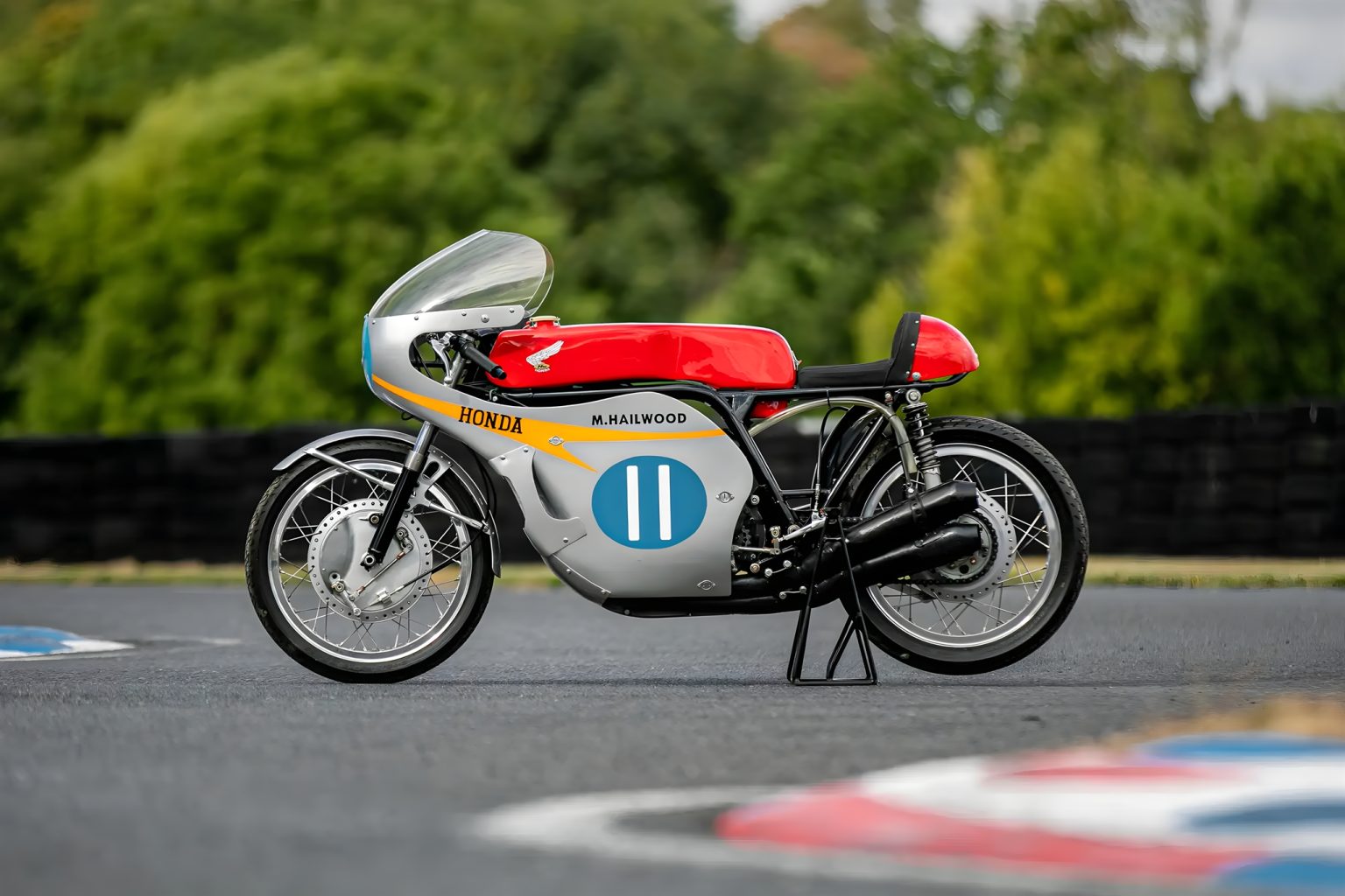 Hailwoods Honda RC173 kommt unter den Hammer: Rekordpreis in Sichtweite Restaurierte Honda RC173 von Mike Hailwood in silbergrauer Werksfarbe mit rotem Tank, orangefarbenem Streifen und Startnummer II, Seitenansicht rechts auf einer Rennstrecke vor grünem Blätterhintergrund