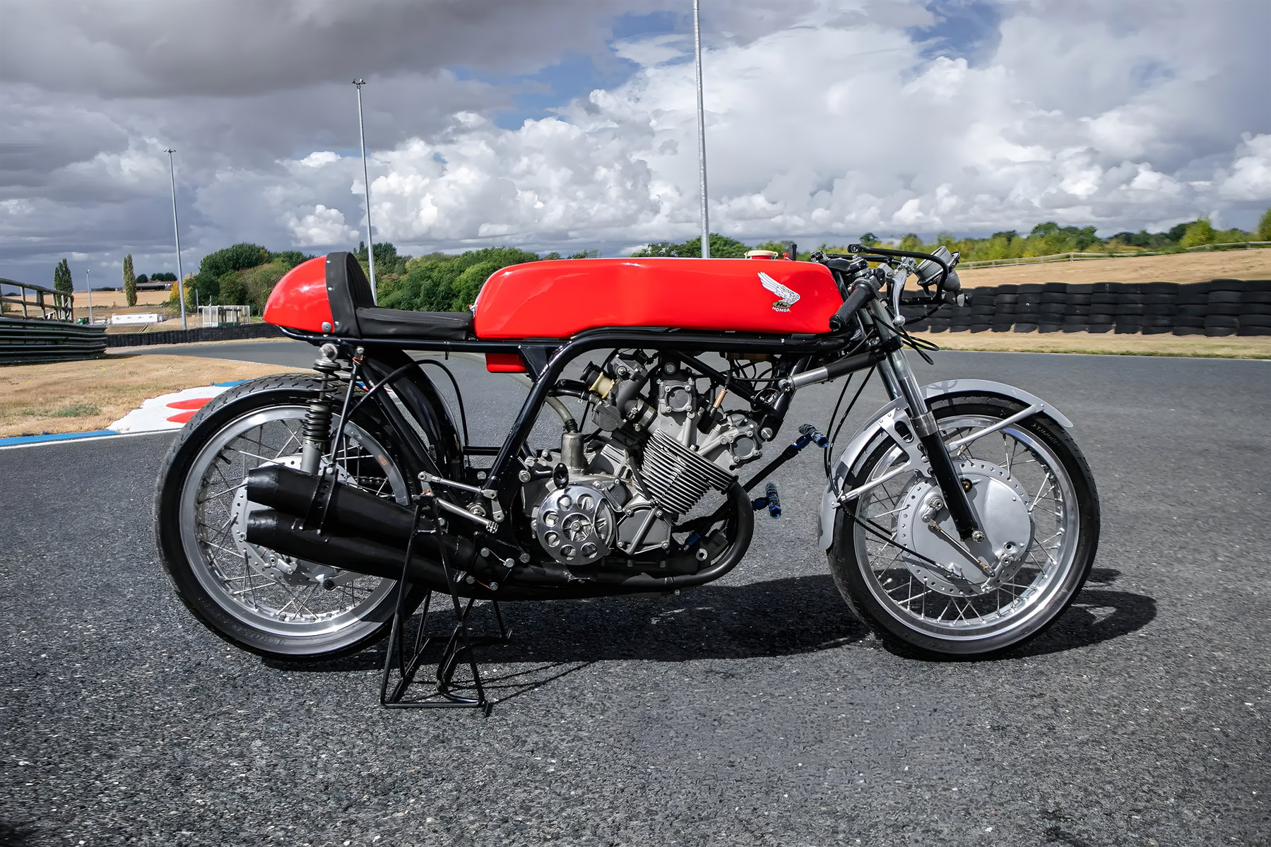 Hailwoods Honda RC173 kommt unter den Hammer: Rekordpreis in Sichtweite 3 Honda RC173 auf Achsständer, Seitenansicht mit Blick auf den freiliegenden luftgekühlten Vierzylindermotor, doppelte Auspuffkrümmer und roten Tank vor bewölktem Himmel