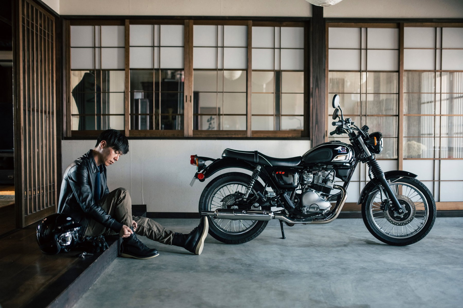 Kawasaki Meguro S1 2027 in Ebony vor einem traditionellen japanischen Haus mit Shoji-Türen, davor ein Fahrer in Lederjacke, der seine Stiefel schnürt
