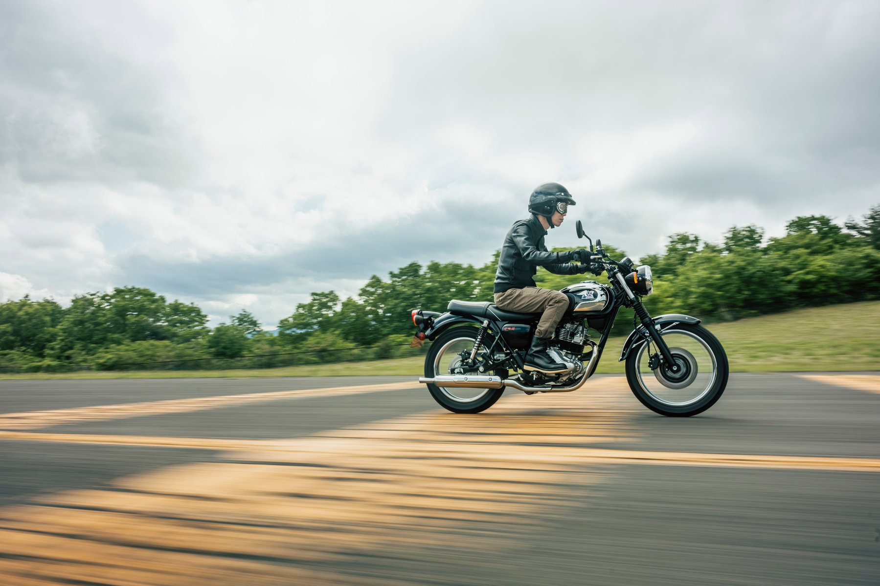 Kawasaki Meguro S1 2027 in Ebony in Fahrt auf einer Landstraße – Seitenansicht mit Fahrer in schwarzer Lederjacke vor grüner Hügellandschaft