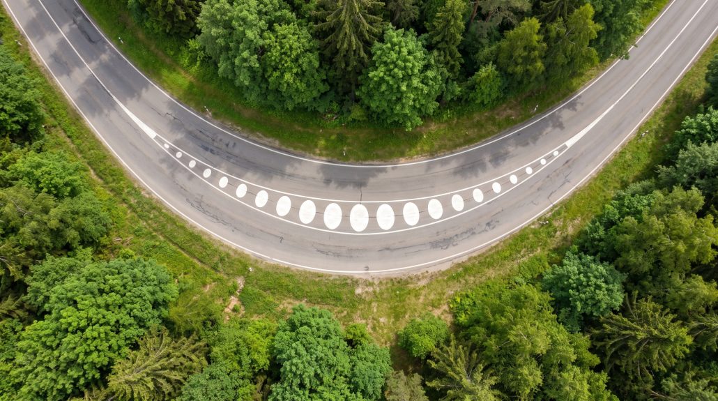Kurvenmarkierungen für Motorräder: Norwegen zieht nach, NRW weitet aus 1 KI-generierte Luftaufnahme einer Linkskurve mit weissen Ellipsen-Fahrbahnmarkierungen entlang der Mittellinie in einer Waldlandschaft