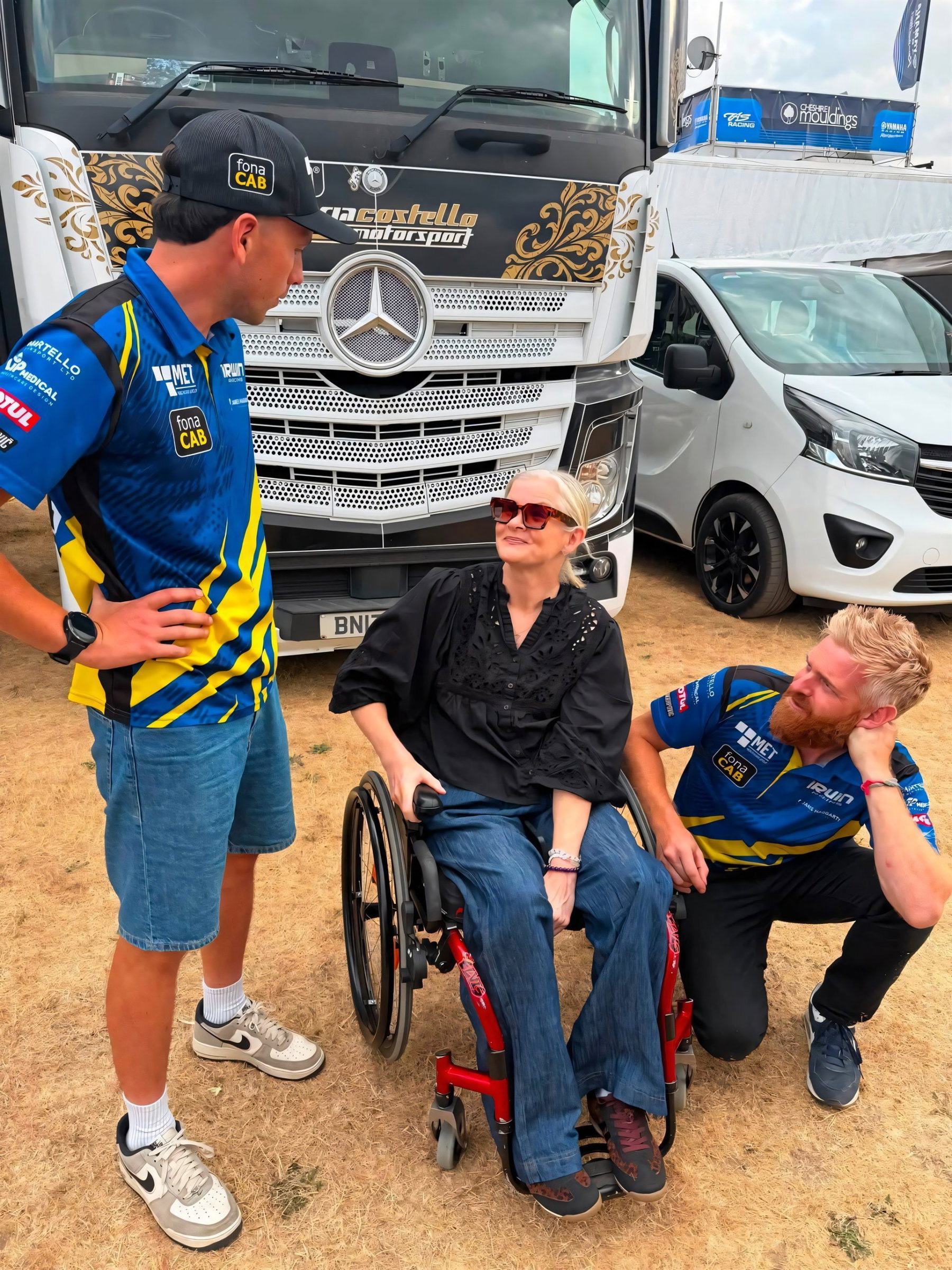 Maria Costello zurück im Fahrerlager: Was der Besuch in Thruxton bedeutet 1 Maria Costello im Rollstuhl im Gespräch mit zwei Teammitgliedern in blau-gelber Irwin-Racing-Teamkleidung vor dem Team-Truck im BSB-Fahrerlager Thruxton