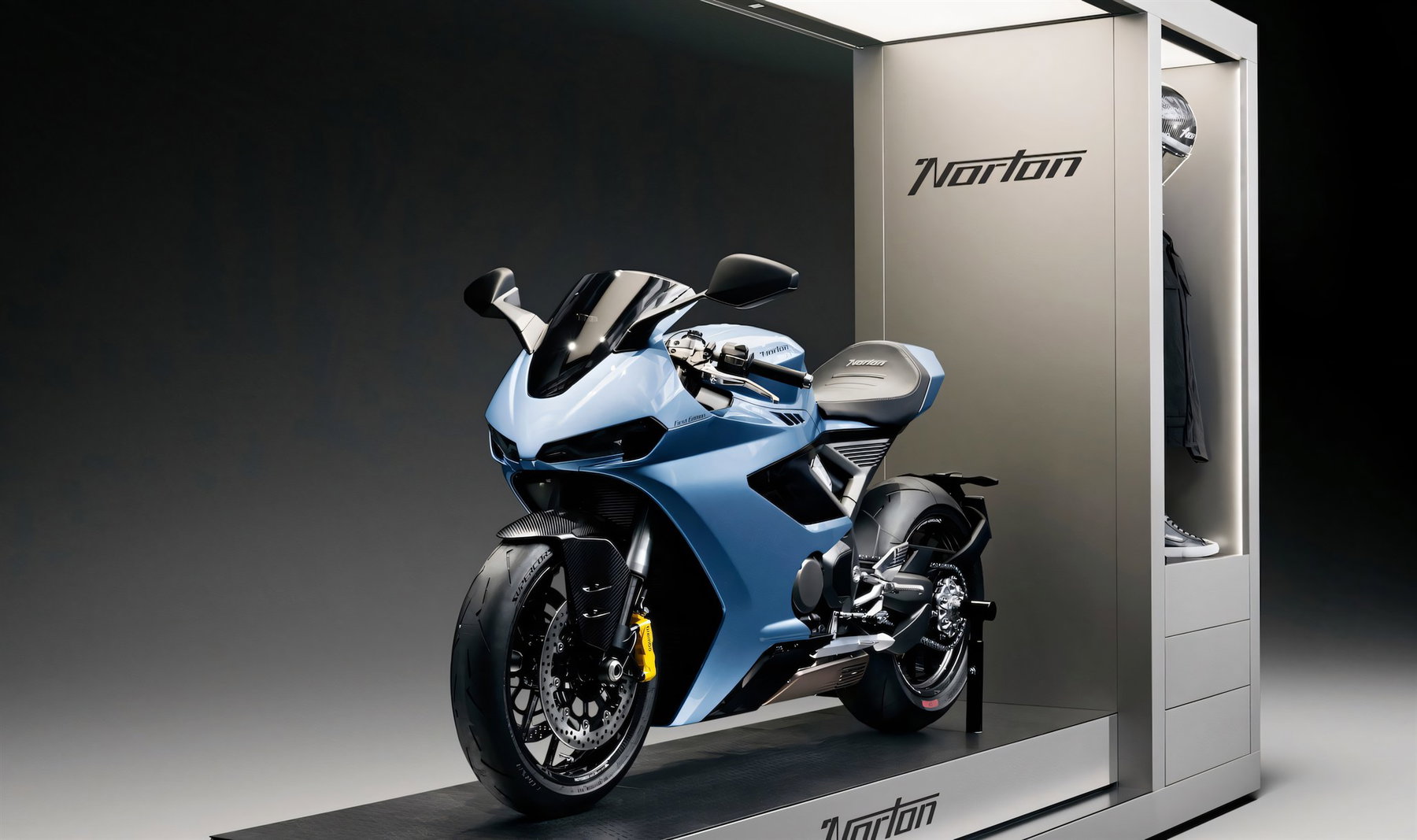Norton overhauls dealer network: showroom concept from 50 square metres 2 Norton Showroom-Konzept (2026) – 3/4-Heckansicht mit Manx-R-Typenschild am Heck, im Hintergrund Vitrinensaeule mit Grafikpanel und Bekleidung