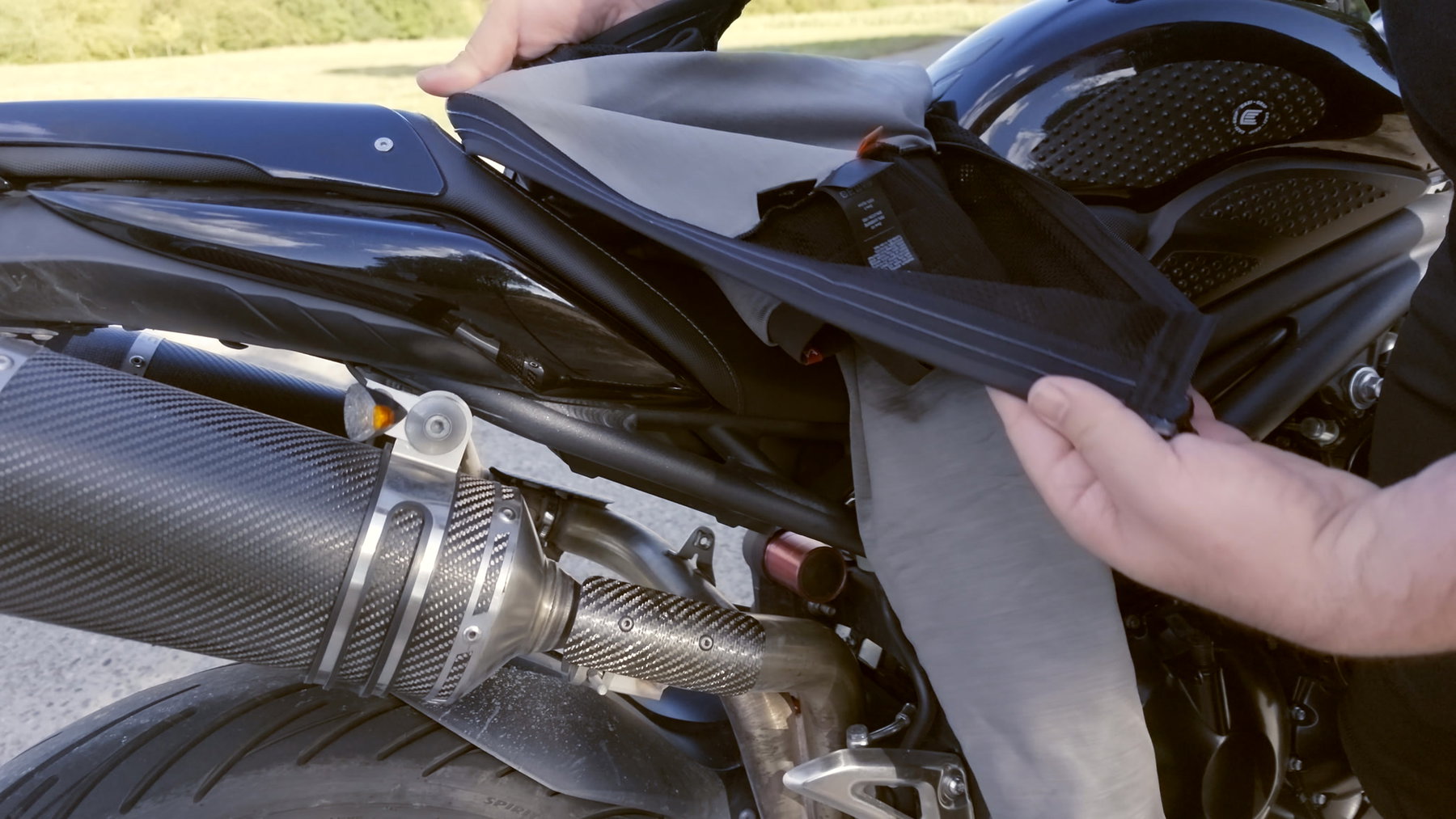Pando Moto Commando UH und Skin UH 03: Schutzkleidung, die niemand sieht 2 Detail der Pando Moto Skin UH 03 – eine Hand hält das Hosenbein mit Verschluss und Nahtverlauf ins Licht, dahinter Heck und Auspuff der Triumph