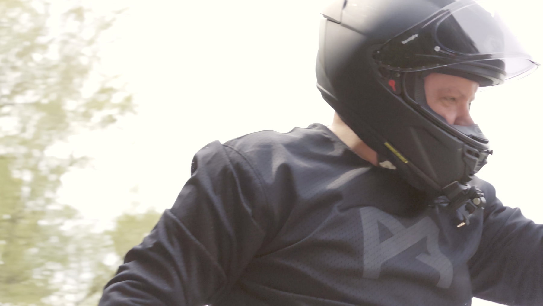 Pando Moto Commando UH und Skin UH 03: Schutzkleidung, die niemand sieht 5 Formatfüllende Nahaufnahme des Fahrers auf der Triumph Speed Triple 1050 – dunkles Pando-Moto-Oberteil mit PM-Brustlogo und schwarzer Integralhelm im Fahrtwind