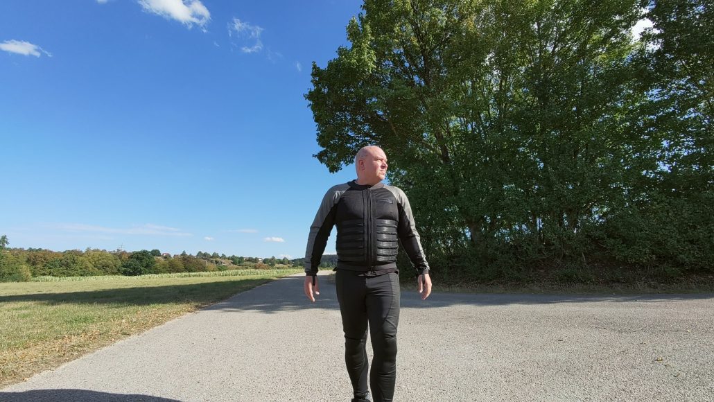 Pando Moto Commando UH und Skin UH 03: Schutzkleidung, die niemand sieht 1 Drohnenaufnahme auf Augenhöhe: Der Fahrer geht in der Pando-Moto-Kombination aus grau-schwarzem Oberteil und enger Hose den Feldweg entlang, links das abgeerntete Feld, rechts eine Baumreihe
