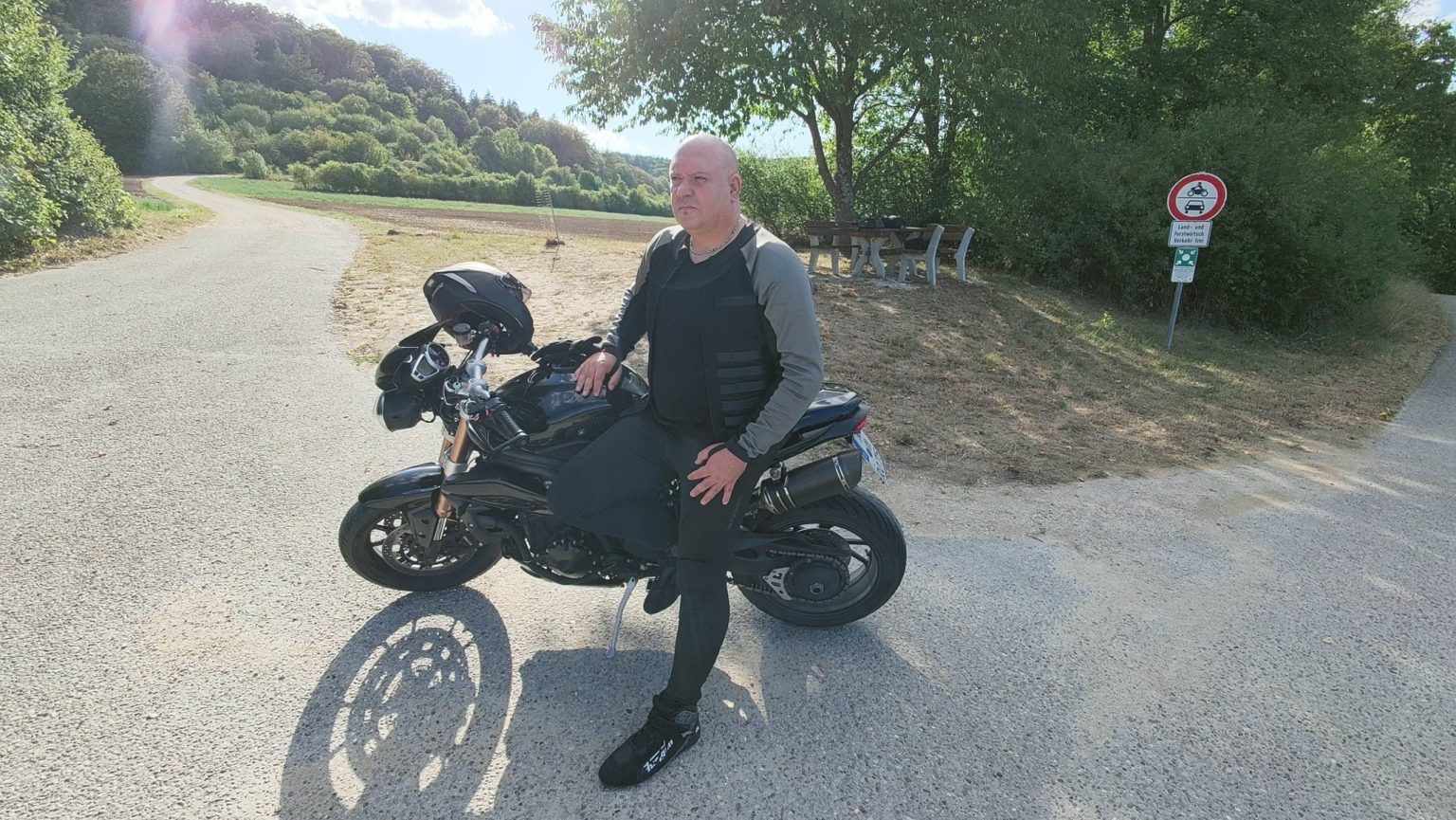 Drohnenaufsicht am späten Nachmittag: der Fahrer lehnt in der Pando-Moto-Kombination an der Triumph Speed Triple 1050, der Schatten von Motorrad und Fahrer fällt lang über den Kiesplatz