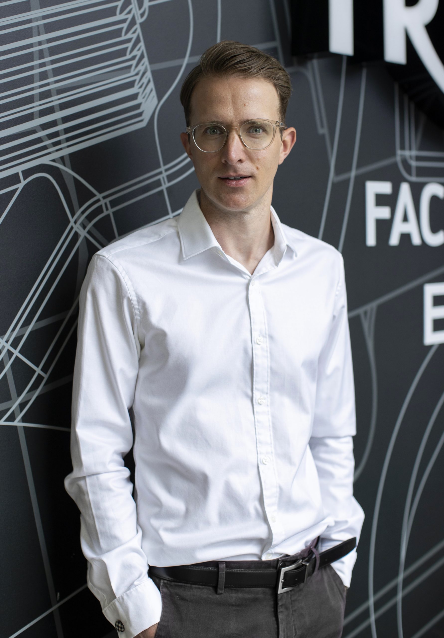 Triumph trennt Vertrieb und Marketing: BMW-UK-Chef George kommt 5 Nick Bell, neuer Chief Marketing Officer von Triumph Motorcycles – Porträt im weißen Hemd mit Brille vor dunkler Triumph-Factory-Wandgrafik