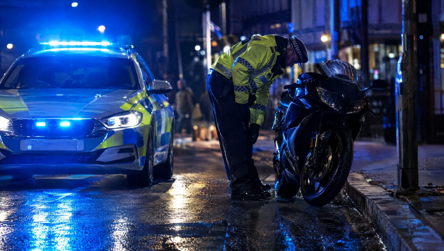 Naechtliche Polizeikontrolle eines Motorrads in der Stadt: Ein Polizeibeamter in Warnweste kontrolliert ein schwarzes Sportmotorrad neben einem Streifenwagen mit blauem Blaulicht auf regennasser Strasse (Symbolbild, KI-generiert)