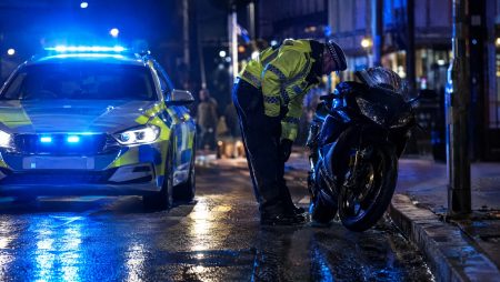 Naechtliche Polizeikontrolle eines Motorrads in der Stadt: Ein Polizeibeamter in Warnweste kontrolliert ein schwarzes Sportmotorrad neben einem Streifenwagen mit blauem Blaulicht auf regennasser Strasse (Symbolbild, KI-generiert)