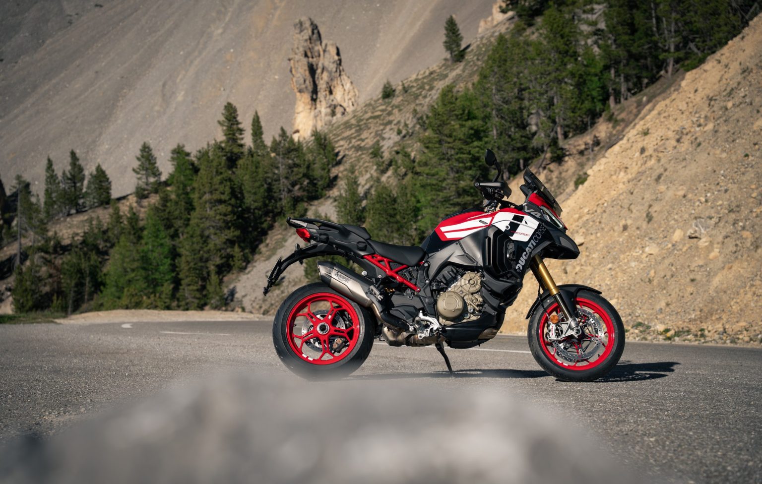 Ducati Multistrada V4 Pikes Peak 2027 in der neuen Ducati-Corse-Lackierung in Rot, Weiß und Schwarz mit roten Schmiederädern, rechte Seitenansicht auf einer Passstraße vor Felsnadeln und Nadelwald