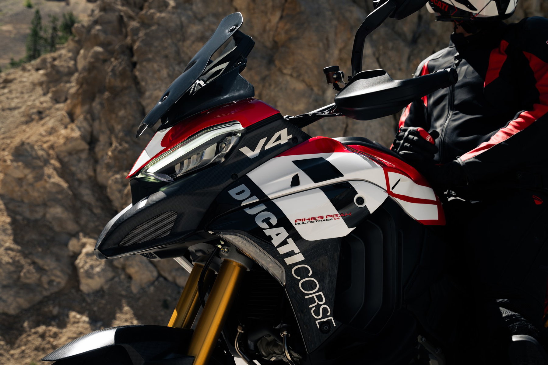 Ducati Multistrada V4 Pikes Peak 2027: MotoGP Look, Same Technology 3 Detailaufnahme der Front der Ducati Multistrada V4 Pikes Peak 2027 mit LED-Scheinwerfer, V4-Logo, Carbon-Schnabel, Ducati-Corse-Schriftzug und goldenen Öhlins-Gabelrohren, daneben ein Fahrer in Ducati-Lederkombi