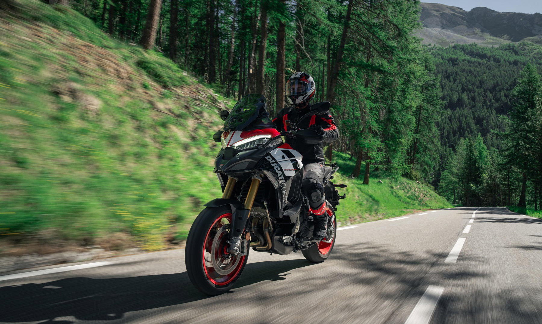 Ducati Multistrada V4 Pikes Peak 2027: MotoGP Look, Same Technology 6 Fahrer auf der Ducati Multistrada V4 Pikes Peak 2027 mit roten Felgen und Ducati-Corse-Lackierung in voller Fahrt auf einer Waldstraße in den Bergen