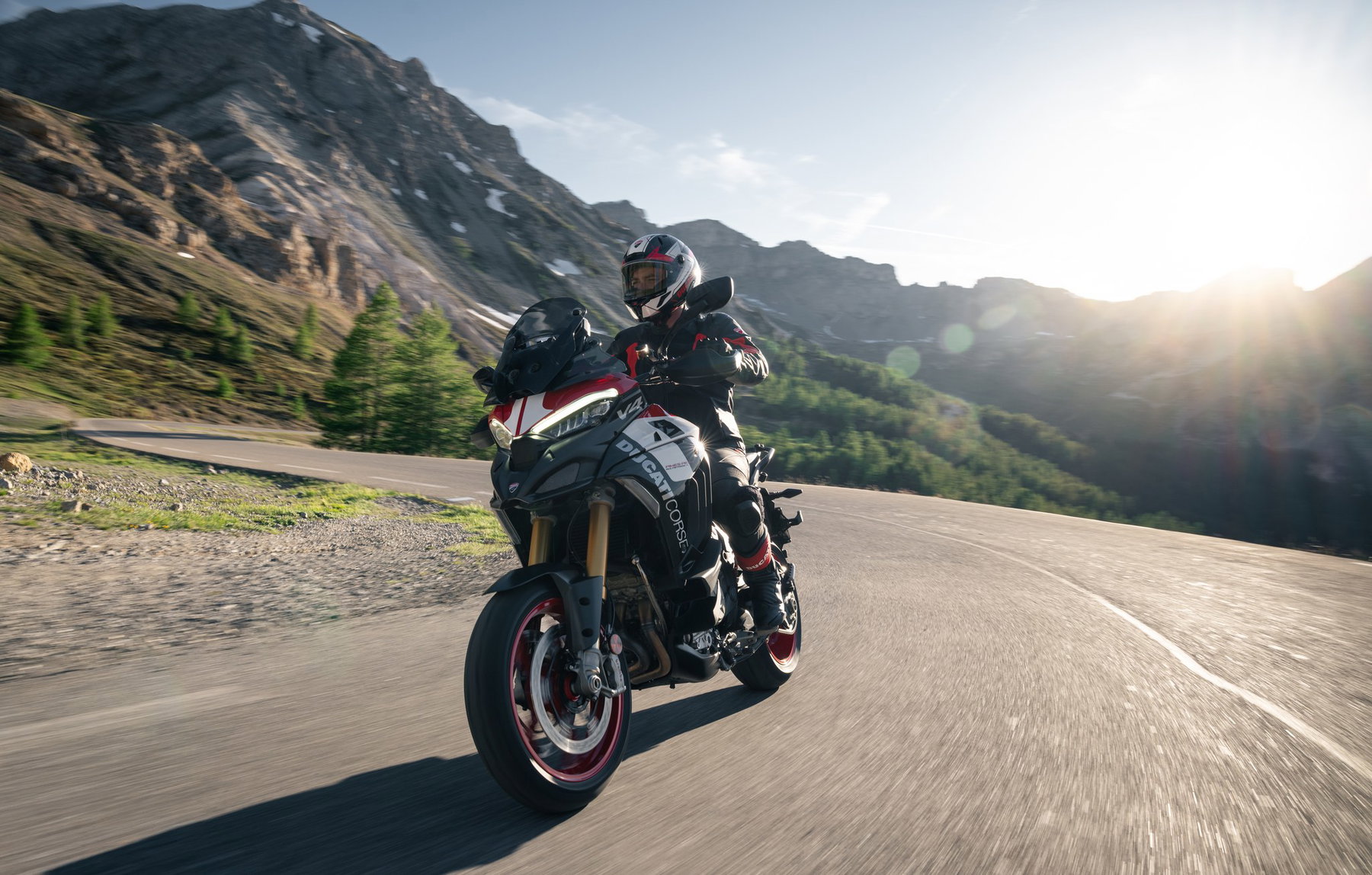 Ducati Multistrada V4 Pikes Peak 2027: MotoGP Look, Same Technology 5 Ducati Multistrada V4 Pikes Peak 2027 mit Fahrer bei Gegenlicht in einer Kurve einer Alpenpassstraße, im Hintergrund Berggipfel mit Schneeresten
