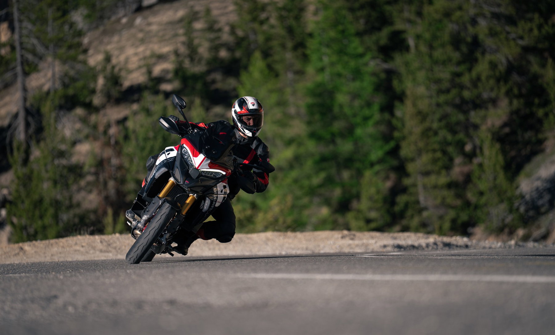 Ducati Multistrada V4 Pikes Peak 2027: MotoGP Look, Same Technology 2 Fahrer mit weiß-rotem Helm legt die Ducati Multistrada V4 Pikes Peak 2027 mit ausgestelltem Knie tief in eine Linkskurve, im Hintergrund ein bewaldeter Hang