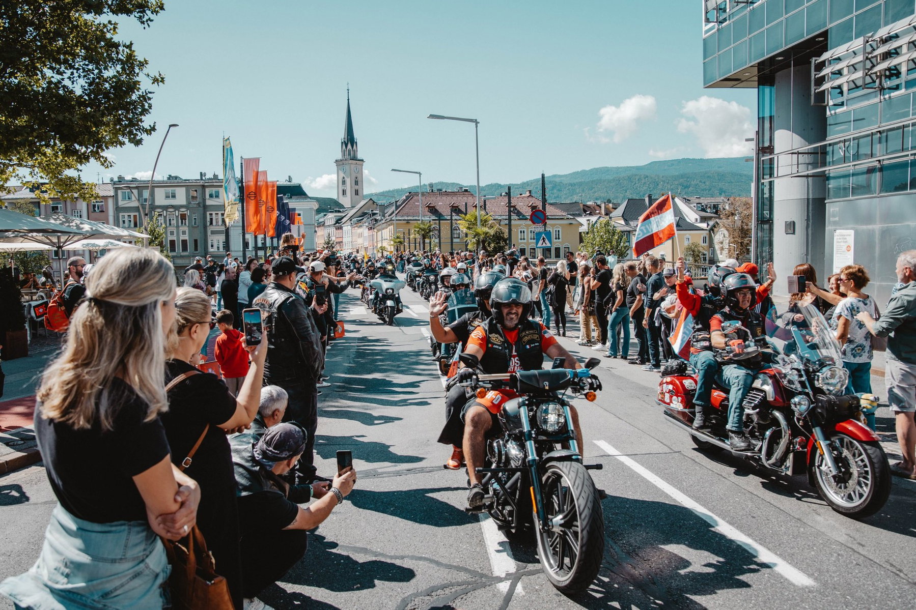 European Bike Week 2026: What Remains of the Harley Rally at Lake Faak 2 European Bike Week 2026 – Harley-Davidson-Parade durch die Villacher Innenstadt, winkende Fahrer vorbei an Zuschauern mit Smartphones, im Hintergrund Kirchturm und Kärntner Berge